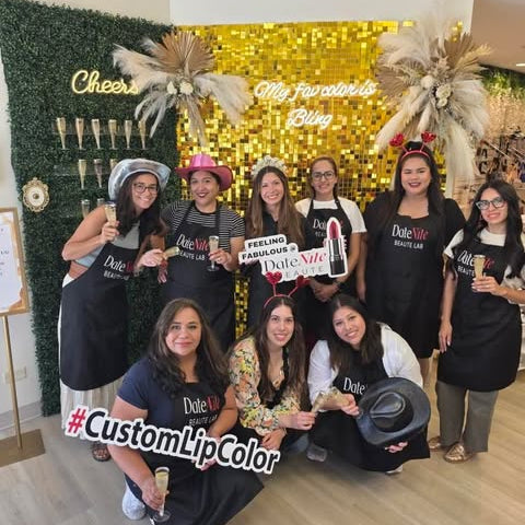 A group of nine women posing together in a decorated indoor space with a gold glitter backdrop, greenery wall, and champagne bottles. Several women are wearing black Dole Lip Color aprons, and one person holds a sign that reads 'Dole Lip Color'.