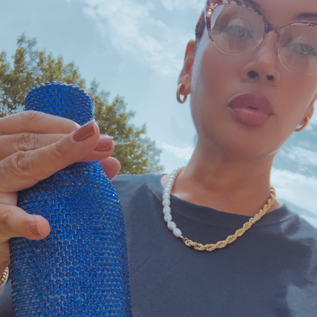 A person outdoors holding a tall, blue rhinestone-covered bottle or tumbler. The person is wearing a dark gray t-shirt, a gold chain necklace, and glasses.