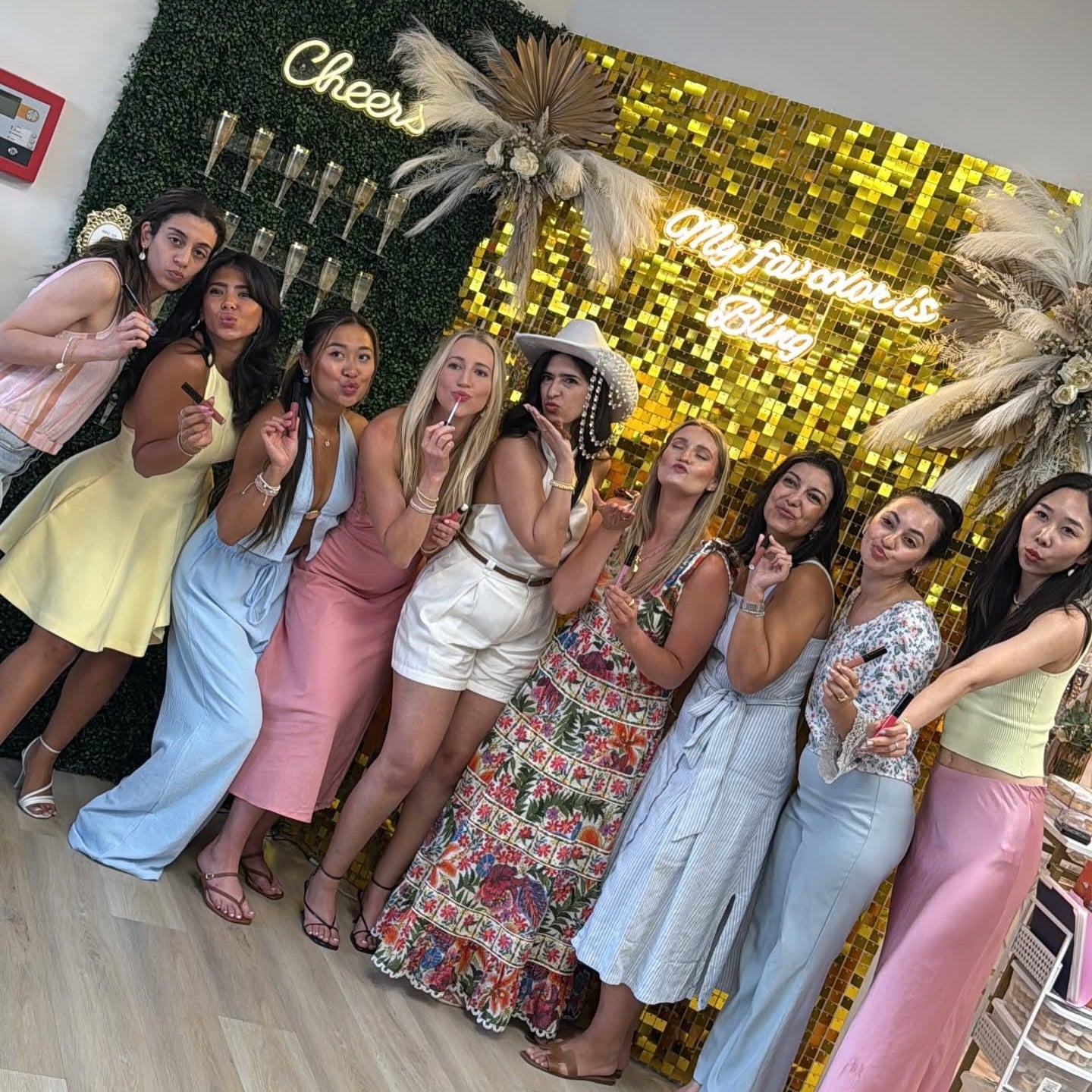 A group of eight women posing together in front of a decorated party backdrop with a green hedge, gold sequin wall, and neon signs reading 'Cheers' and 'We have a great time!'.