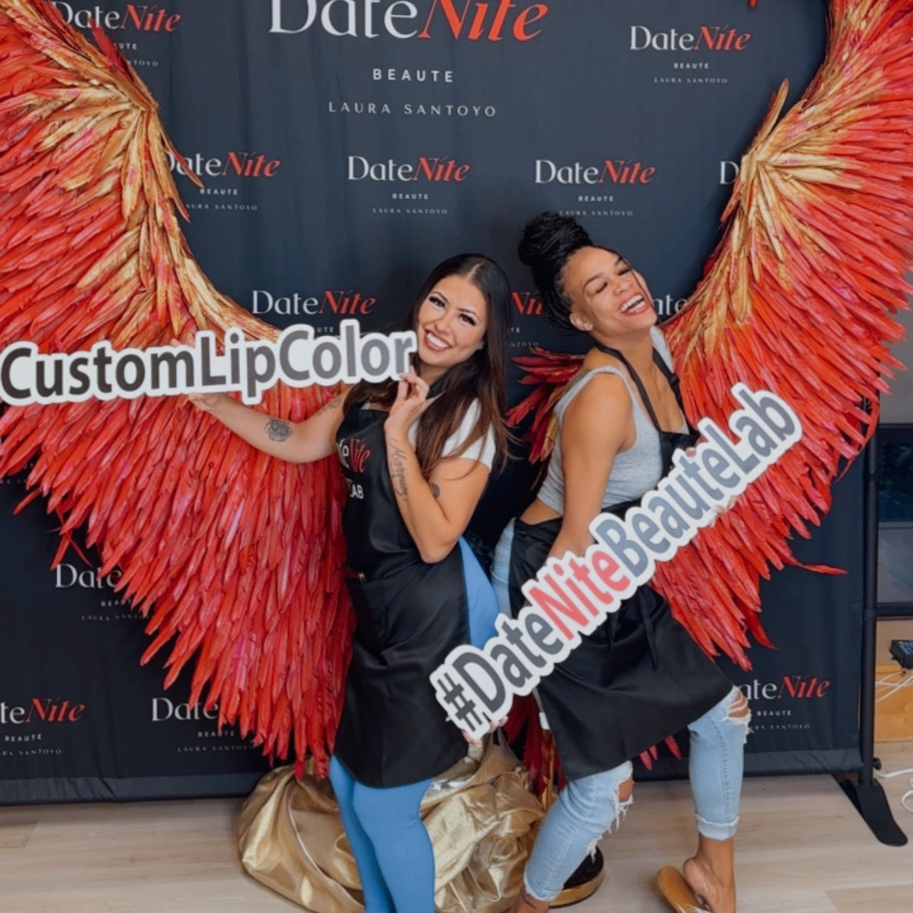 Two women posing in front of a black DateNite backdrop with large red feather wings on either side, holding a white sign that reads #DateNiteBeautyLab. A banner on the left says CustomLipColor.