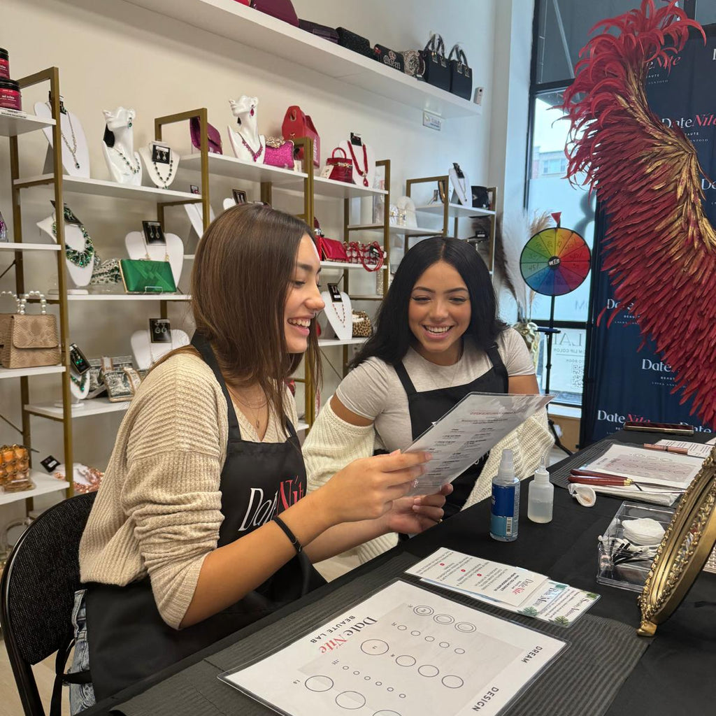 Two women wearing black aprons smile and laugh behind a counter in a boutique, holding a printed sheet. Shelves in the background display various handbags and accessories, and a large red feather decoration is visible on the right.