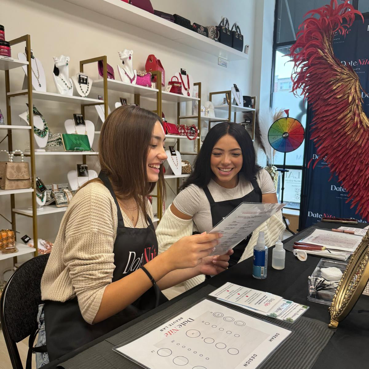 Two women wearing black aprons smile and laugh behind a counter in a boutique, holding a printed sheet. Shelves in the background display various handbags and accessories, and a large red feather decoration is visible on the right.