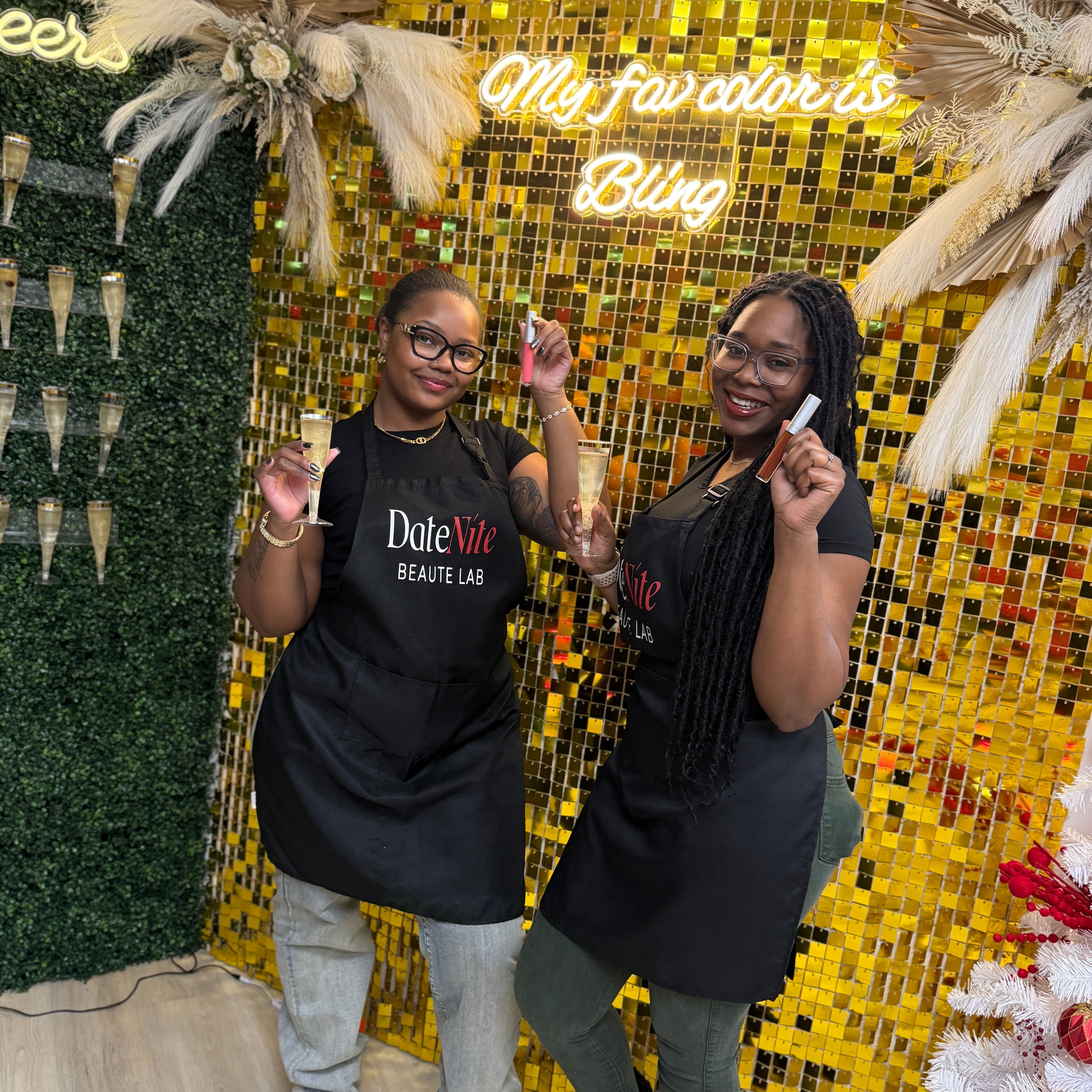 Two women wearing black DuteeV2 Beauty Lab aprons pose in front of a gold mosaic wall with decorative dried grasses and a neon sign that reads 'My fav color is Bling'.